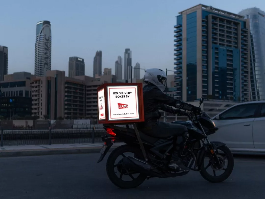 Motorcycle with a lit LED delivery box ad passing the Dubai skyline at dusk, moving digital OOH advertising in Dubai
